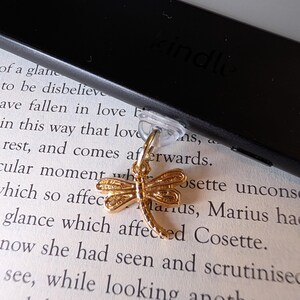 May include: A gold-toned metal dragonfly charm attached to a clear plastic clip. The charm features textured wings and a segmented body. The clip is secured to the edge of a black electronic reading device that rests on a page of printed text.