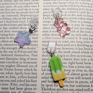 May include: Three clear plastic page markers, each featuring a dangling charm: a glittery purple star, a translucent pink gummy bear, and a green and yellow popsicle. The markers are displayed against a background of printed book text.