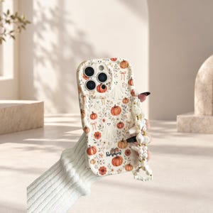 Peut inclure: Une coque de téléphone couleur crème avec un motif répétitif de citrouilles orange, de petits fantômes et de nœuds, avec le texte Pumpkins imprimé près du bas. La coque comprend une dragonne perlée décorative avec des accents blancs et dorés.