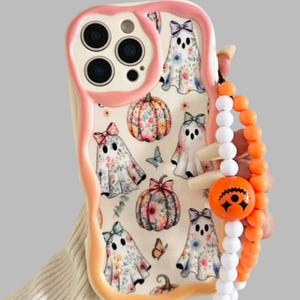 May include: A cream-colored phone case with a wavy pink border featuring a pattern of ghosts wearing bows and floral pumpkins. A beaded phone charm with white and orange beads and a round orange jack-o'-lantern face bead is attached to the side.