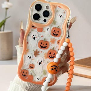 Peut inclure: Une coque de téléphone transparente avec un bord orange ondulé ornée d'un motif de fantômes blancs, de citrouilles d'Halloween orange et de feuilles d'automne. Elle comprend une dragonne perlée avec des perles blanches, orange et une breloque citrouille.