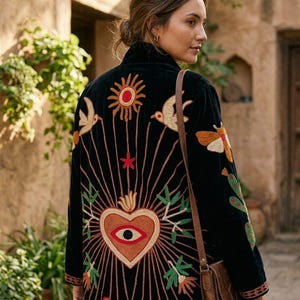 May include: A black velvet long coat featuring intricate embroidery on the back. The design includes a central heart with an eye, a sun, birds, insects, and the word LOVE in capital letters. The coat has long sleeves and a brown leather shoulder bag.