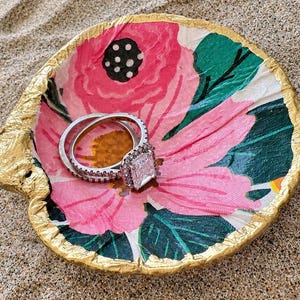 May include: A decorative seashell trinket dish featuring a decoupage floral pattern with pink petals, green leaves, and a gold-painted rim. Two silver-toned rings with clear stones are resting inside the shell, which has a textured, organic shape.