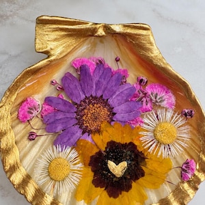 May include: A decorative seashell dish with a gold-painted rim and interior, featuring a collection of pressed dried flowers embedded in a clear resin layer. The arrangement includes a large purple daisy, a yellow sunflower, and small white daisies.