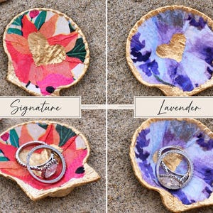 May include: Two decorative seashell dishes with gold-painted edges. The left dish features a floral pattern with a gold heart and the text Signature. The right dish has a purple floral pattern with a gold heart and the text Lavender. Both hold rings.