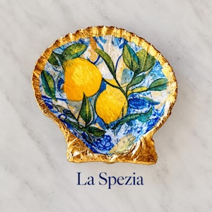 May include: A decorative seashell dish featuring a vibrant print of two yellow lemons with green leaves against a blue and white patterned background. The scalloped edges of the shell are finished with a metallic gold trim. The text La Spezia is below.