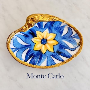 May include: A decorative shell dish featuring a central yellow flower motif surrounded by blue and white floral patterns, with a metallic gold painted rim. The text Monte Carlo is printed in dark blue below the shell-shaped trinket tray.