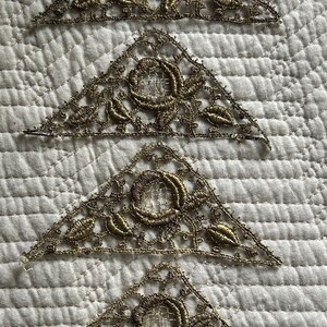 May include: Four gold-coloured lace triangular appliques with intricate floral designs. The appliques are arranged in a stack, with the largest at the bottom and the smallest at the top.