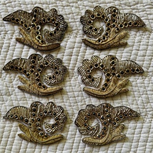 May include: Six gold and silver embroidered floral appliques with black beads. The appliques are in the shape of a stylised flower with a central circle and a curved stem.