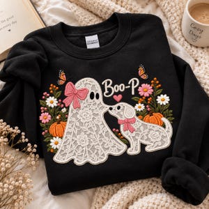 Könnte beinhalten: Ein schwarzes Sweatshirt mit Rundhalsausschnitt und einer Stickerei, die ein weißes Spitzen-Gespenst und einen weißen Spitzen-Hund zeigt. Das Design umfasst rosa Schleifen, orangefarbene Kürbisse, bunte Blumen, Schmetterlinge und den Text Boo-P.