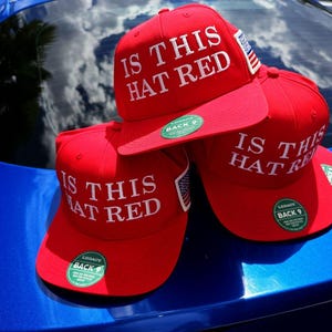May include: Three bright red baseball caps with flat brims, each featuring the white embroidered text IS THIS HAT RED on the front. Each cap has a small American flag patch on the side and a circular green sticker on the brim that reads LEGACY BACK 9.
