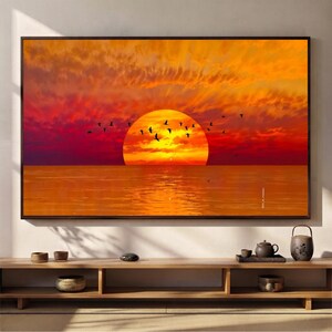 Op de afbeelding: Een rechthoekige wandkunstprint met een levendige zonsondergang boven een kalme oceaan. De grote, felgele zon staat aan de horizon, omringd door dieporanje en rode wolken. Een zwerm zwarte vogels is afgebeeld tegen de gloeiende lucht.