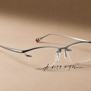 Puede incluir: Gafas sin montura con armazón de metal oscuro, con varillas delgadas que terminan en pequeños detalles circulares rojos. El puente y las almohadillas nasales están integrados en la estructura. El texto japonés "かわさきかずお" es visible.
