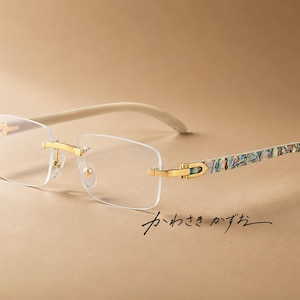 Puede incluir: Gafas sin montura con lentes rectangulares transparentes, puente y bisagras de metal dorado, y patillas con un patrón iridiscente multicolor similar a una concha. Las puntas de las patillas son de color crema. El texto japonés "かわさき かずお" es visible.