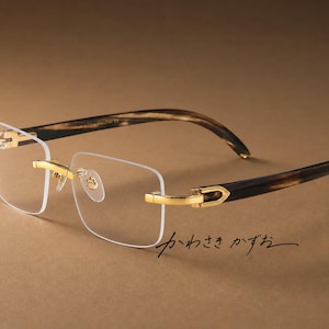 Puede incluir: Gafas sin montura con lentes rectangulares transparentes, puente y bisagras de metal dorado y patillas con patrón de carey marrón oscuro. El texto japonés "かわさき かずお" es visible en escritura negra debajo del marco.