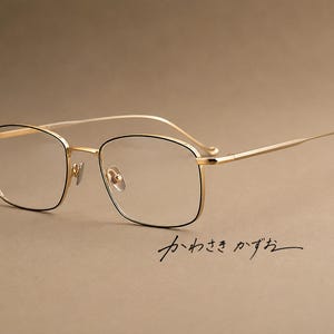 May include: Gold-toned metal eyeglasses featuring a rectangular frame with thin black rims around the lenses, slender temples, and adjustable nose pads. The Japanese text "かわさき かずお" is visible below the frame on the right side.