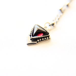 May include: A silver-toned metal pendant featuring a triangular shape with a dark, polished stone center and a deep red accent. The pendant is attached to a delicate silver-toned chain, showcasing intricate metalwork details along the edges.