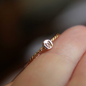May include: A gold ring with a twisted band and a small round charm with the letter "M" engraved on it.
