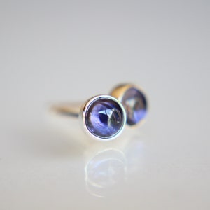 Peut inclure: Une paire de boucles d'oreilles à clous avec des pierres précieuses rondes, violet foncé, serties dans du métal argenté. Les pierres précieuses ont une qualité réfléchissante, présentées sur une surface blanche.