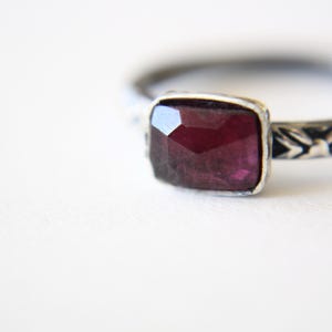 May include: A silver ring featuring a rectangular, faceted deep red garnet gemstone set in a bezel. The metal band displays a dark, oxidized finish with a repeating engraved pattern along the sides, providing a textured appearance to the silver.