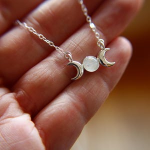 Può includere: Una collana d'argento con un ciondolo di pietra di luna e tre ciondoli a forma di luna crescente. La collana è su una mano.