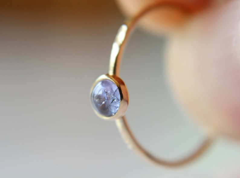 Puede incluir: Un delicado anillo de oro con una sola piedra preciosa redonda de color p&uacute;rpura claro engastada en el centro.