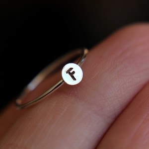 May include: A silver ring with a small round disc featuring the letter "F" engraved on it.