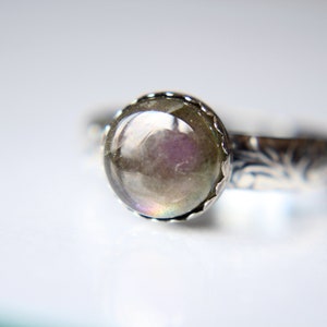 May include: A silver ring with a round, iridescent gemstone set in a scalloped bezel. The band is decorated with a floral design.