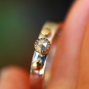 May include: A silver ring with a gray diamond and gold accents. The ring is set with a single, round diamond in the center, surrounded by a gold bezel. The band of the ring is silver and has a textured finish. There are three gold accents on the band, each with a round shape.