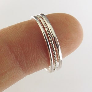 K&ouml;nnte beinhalten: Drei silberne Ringe zum Stapeln, einer mit einem gedrehten Design, an einem Finger getragen.