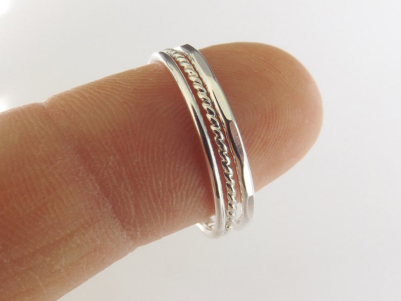 K&ouml;nnte beinhalten: Drei silberne Ringe zum Stapeln, einer mit einem gedrehten Design, an einem Finger getragen.