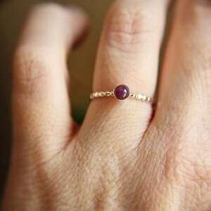 Puede incluir: Un delicado anillo de oro con una peque&ntilde;a piedra preciosa redonda de color p&uacute;rpura oscuro engastada en el centro. El anillo est&aacute; en el dedo de una persona.