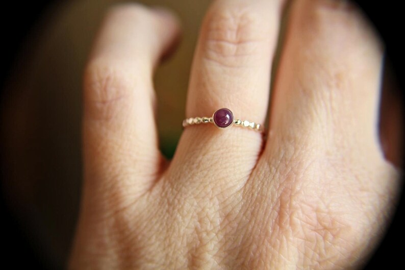 Puede incluir: Un delicado anillo de oro con una peque&ntilde;a piedra preciosa redonda de color p&uacute;rpura oscuro engastada en el centro. El anillo est&aacute; en el dedo de una persona.