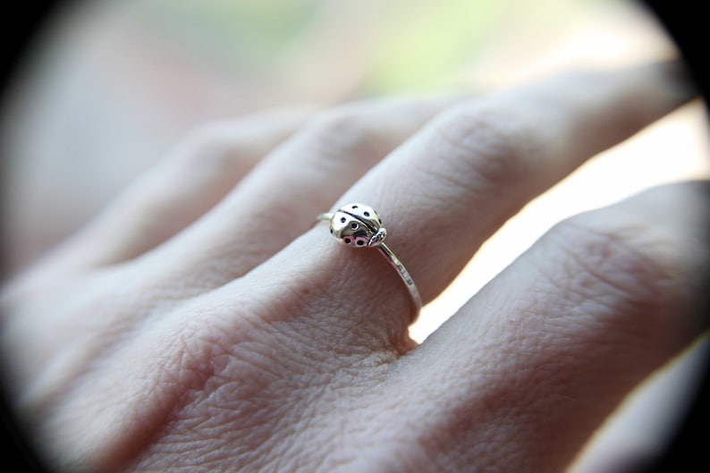 K&ouml;nnte beinhalten: Ein silberner Ring mit einem Marienk&auml;fer-Design. Der Ring wird an einem Finger getragen.