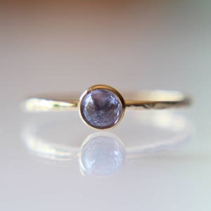 Puede incluir: Un delicado anillo de oro con una sola piedra preciosa redonda de color p&uacute;rpura claro engastada en el centro.