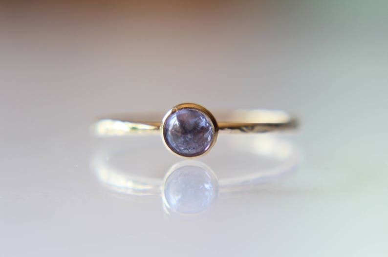 Puede incluir: Un delicado anillo de oro con una sola piedra preciosa redonda de color p&uacute;rpura claro engastada en el centro.