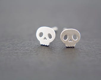 May include: A pair of silver-toned stud earrings shaped like small skulls. Each earring features a smooth, matte finish with cut-out details for the eye sockets, a triangular nose cavity, and a simple row of teeth along the bottom edge.