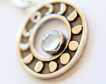 May include: A silver pendant with a moonstone in the center. The pendant is circular and has a design of small circles around the edge.