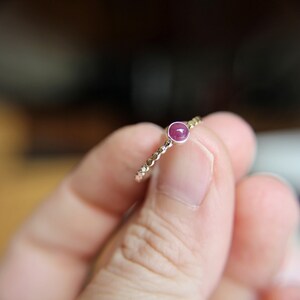 Puede incluir: Un delicado anillo de oro con una peque&ntilde;a piedra preciosa redonda de color rosa engastada en el centro. El anillo est&aacute; en un dedo.