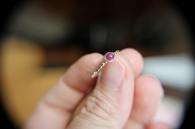 Puede incluir: Un delicado anillo de oro con una peque&ntilde;a piedra preciosa redonda de color rosa engastada en el centro. El anillo est&aacute; en un dedo.