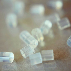 May include: A close-up of a group of small, clear, plastic earplugs scattered on a light brown surface.
