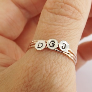 Könnte beinhalten: Drei dünne goldene Ringe mit einem silbernen Buchstaben-Anhänger an jedem. Die Buchstaben sind D, G und J.