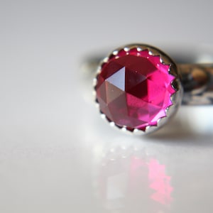 Può includere: Un anello d'argento con una pietra preziosa rosa brillante incastonata in una cornice decorativa a forma di corona.