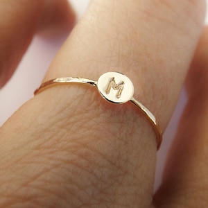 May include: A gold ring with a hammered finish and a round disc featuring the letter "M" engraved on it.