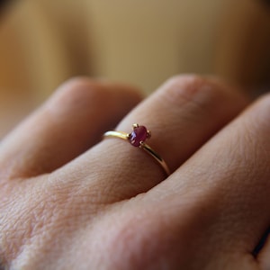 May include: A delicate gold ring with a small, oval-shaped ruby gemstone set in a prong setting.