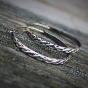 May include: Two silver hoop earrings with a braided design. The earrings are on a wooden surface.