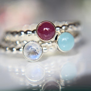 May include: Three silver rings with different colored gemstones. The rings are stacked on top of each other. The top ring has a light blue gemstone, the middle ring has a dark red gemstone, and the bottom ring has a white gemstone.