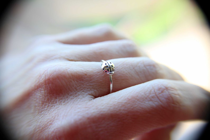 K&ouml;nnte beinhalten: Ein silberner Ring mit einem Marienk&auml;fer-Design. Der Ring wird an einem Finger getragen.