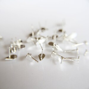 Pu&ograve; includere: Un mucchio di perni per orecchini in argento, sparsi su una superficie bianca. I perni sono progettati per essere utilizzati nella creazione di gioielli.