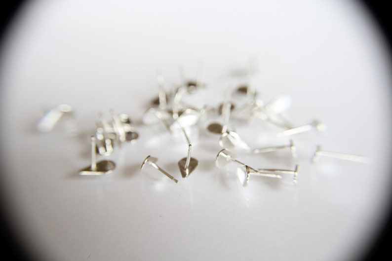 Pu&ograve; includere: Un mucchio di perni per orecchini in argento, sparsi su una superficie bianca. I perni sono progettati per essere utilizzati nella creazione di gioielli.